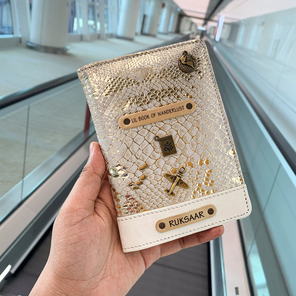A hand holding a white personalized passport cover with gold details and a name tag that reads 'Book of Wanderlust'.