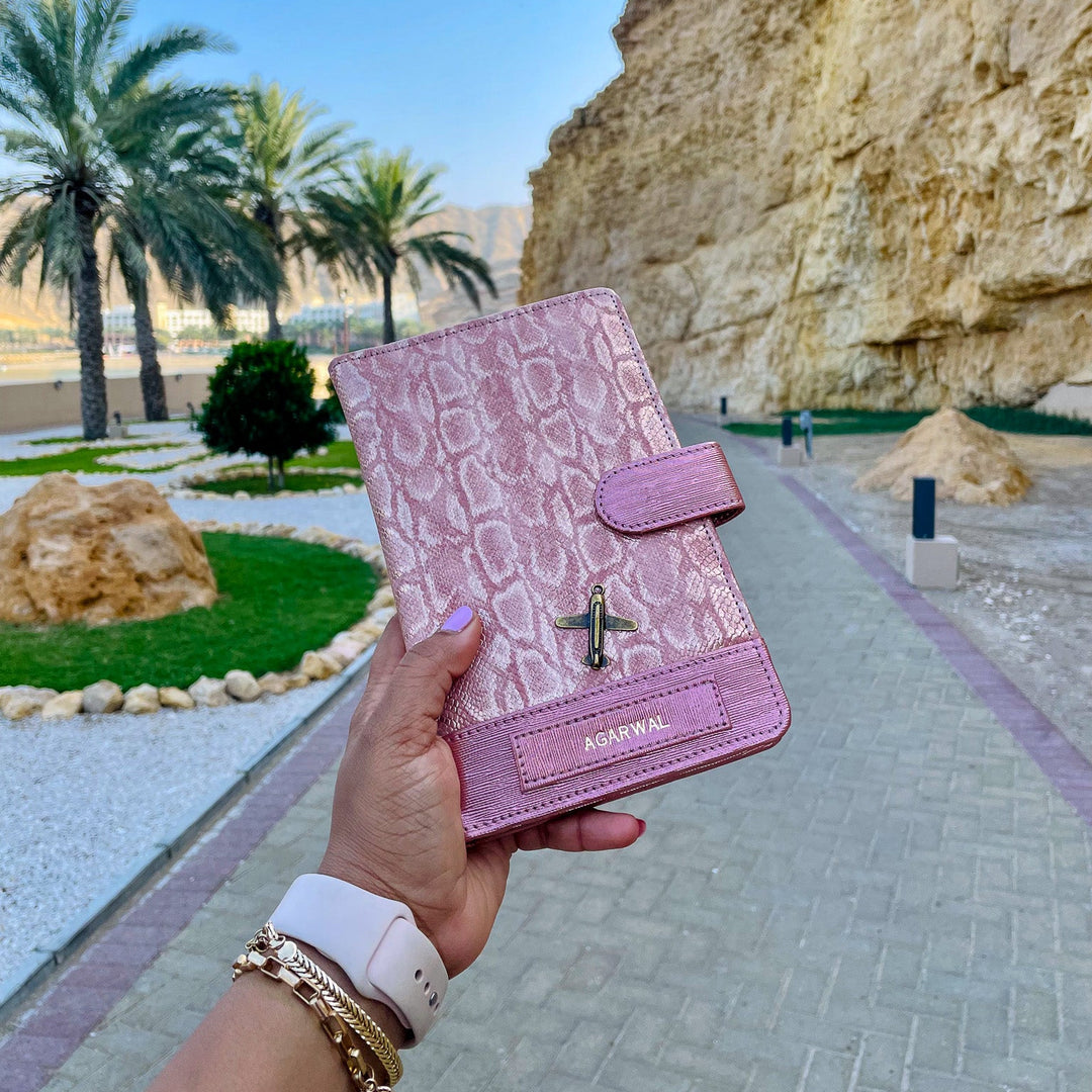 Pink crocodile-patterned wallet held by a hand with a scenic background of palm trees and rocky cliffs.