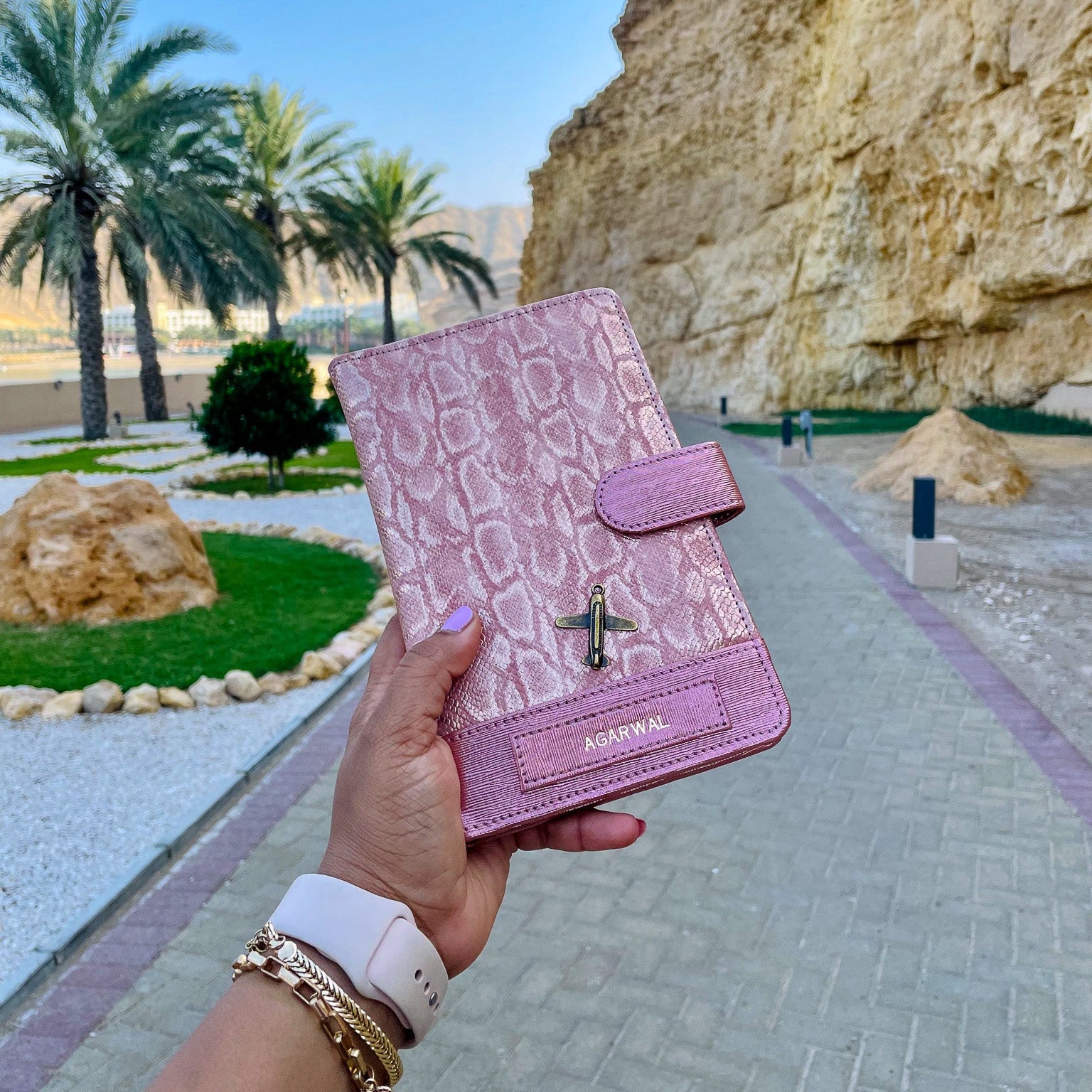 Pink crocodile-patterned wallet held by a hand with a scenic background of palm trees and rocky cliffs.