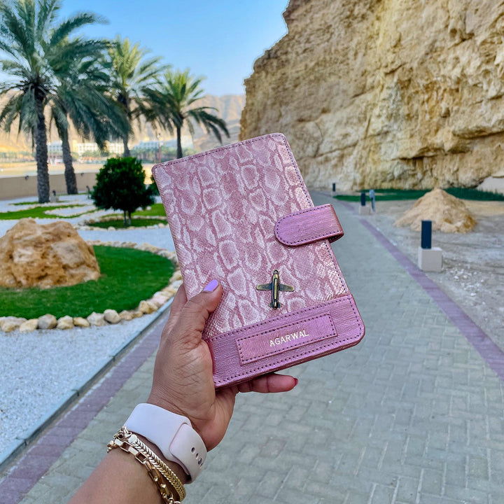Pink crocodile-patterned wallet held by a hand with a scenic background of palm trees and rocky cliffs.