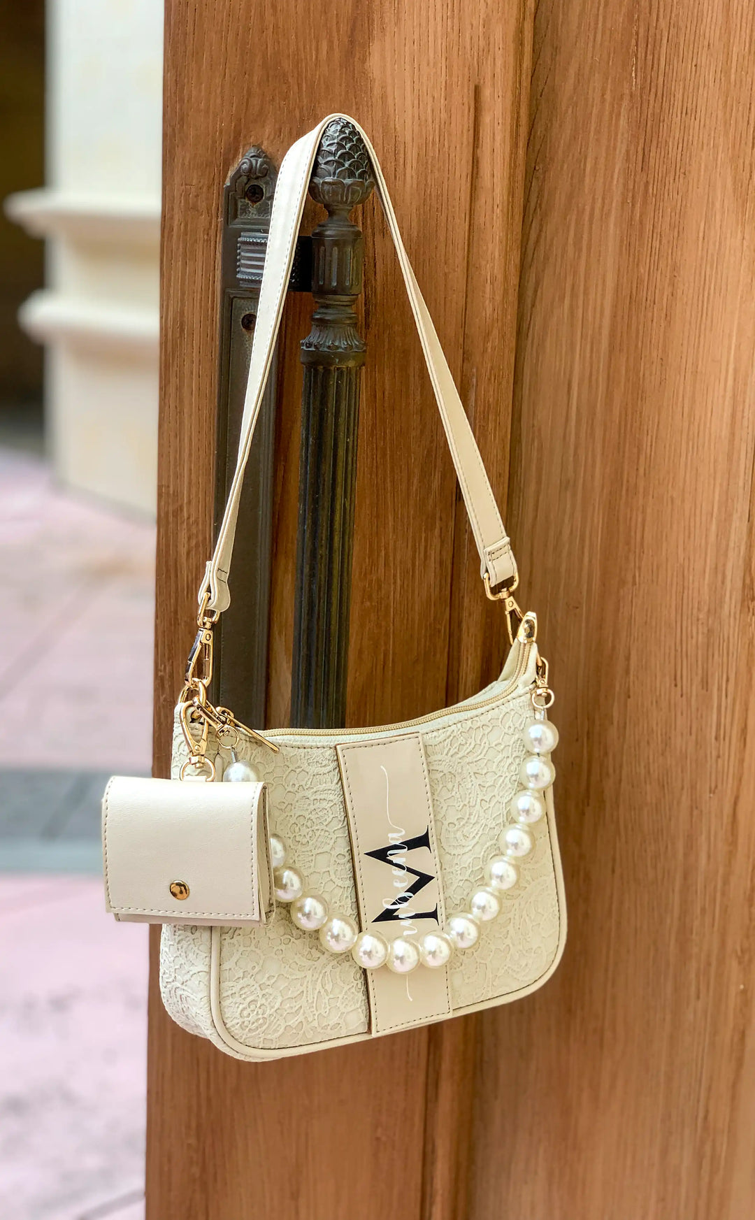 A white mini sling bag with a pearl strap hanging on a wooden surface, showcasing its design and compact size.