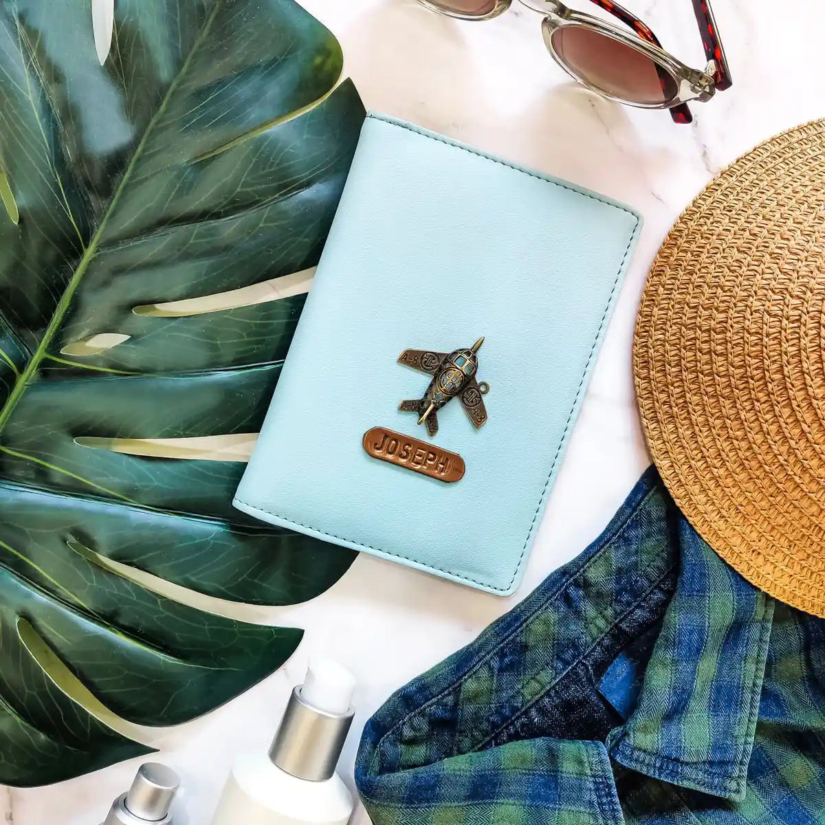 A light blue leather passport holder with a metal airplane charm and a personalized name plate reading 'JOSEPH', styled in a flat lay with a green monstera leaf, sunglasses, a straw hat, blue plaid shirt, and a small bottle on a white marble background - TPC Gifts 