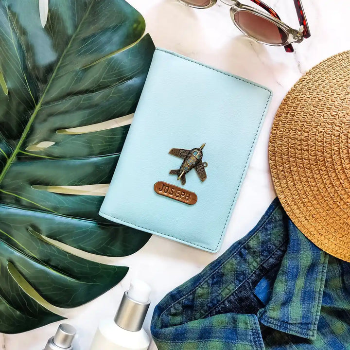 A light blue leather passport holder with a metal airplane charm and a personalized name plate reading &