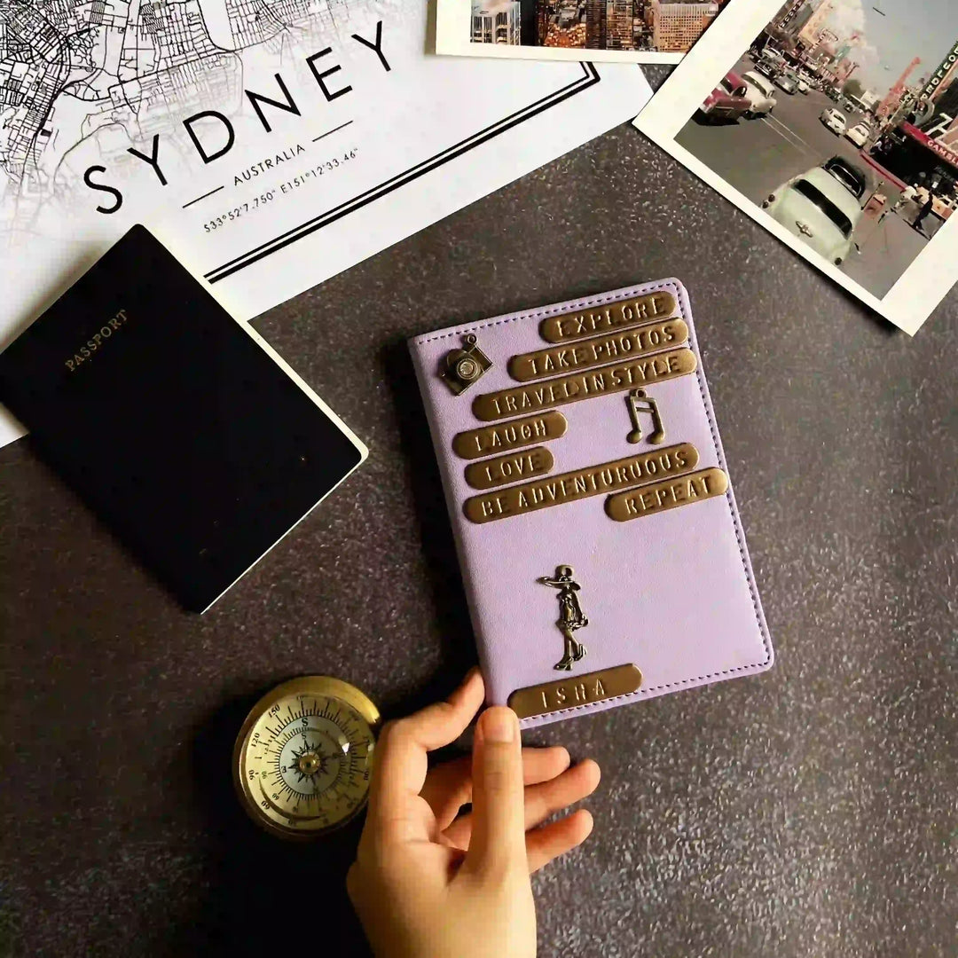 A lavender purple passport cover with gold embossed text and travel-themed designs, held by a hand on a dark tabletop. A black Indian passport, a vintage-style compass, and travel photos and a Sydney map print are arranged around it.
