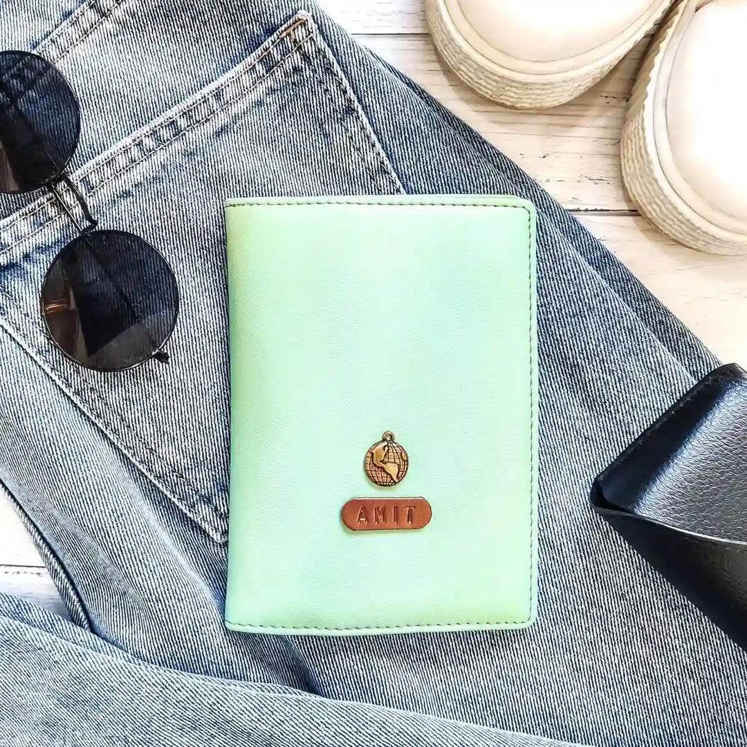 A light green passport cover with a name tag and charm placed on denim fabric. Accessories like sunglasses and shoes are also visible in the background. - TPC Gifts