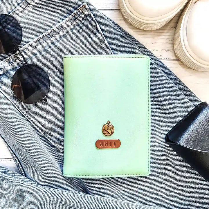 A light green passport cover with a name tag and charm placed on denim fabric. Accessories like sunglasses and shoes are also visible in the background. - TPC Gifts
