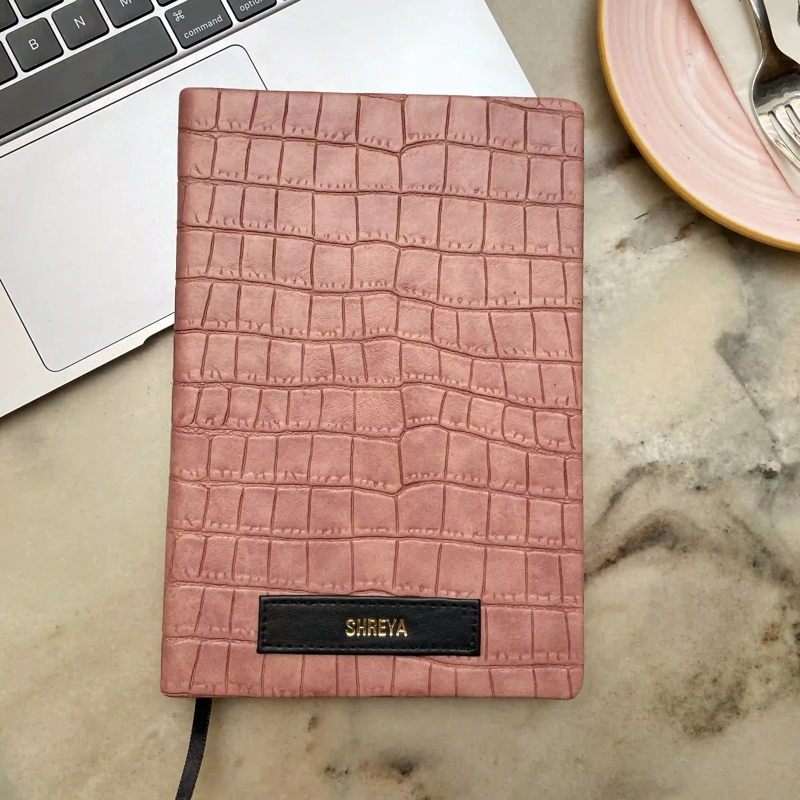 Onion pink crocodile patterned customized diary with a black label, placed on a marble surface next to a laptop and a plate -TPC Gifts