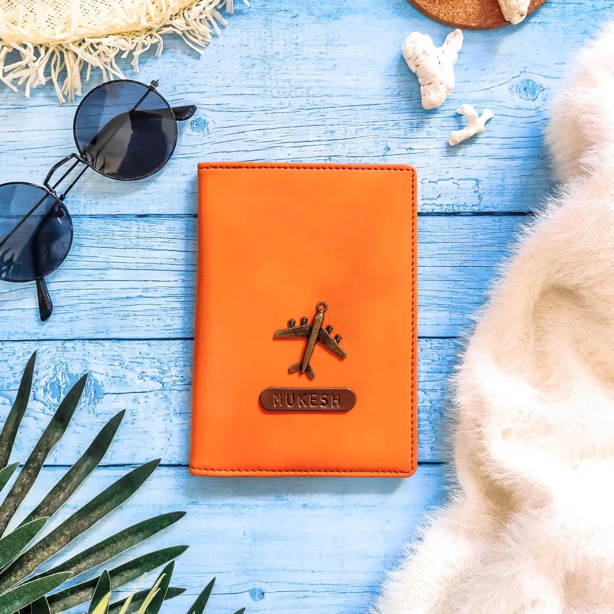 An orange leather passport cover with a metallic airplane emblem and a custom nameplate reading 'NUKESH', styled in a flat-lay on a light blue wooden background with sunglasses, a woven hat, a piece of fruit, and a white faux fur throw.