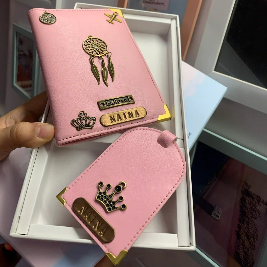A pink personalized card holder wallet and matching luggage tag set held by a hand over an open white box. The set features gold-tone metal corner protectors, a dreamcatcher charm, a crown emblem, and custom nameplates reading &