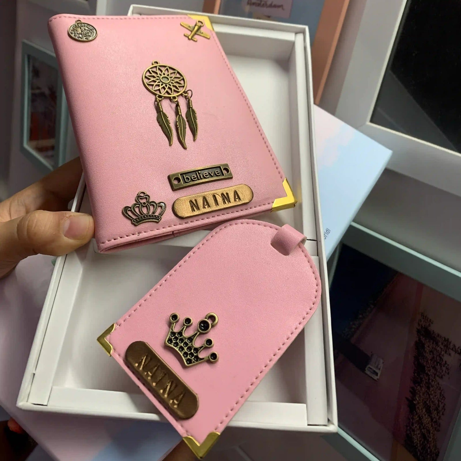 A pink personalized card holder wallet and matching luggage tag set held by a hand over an open white box. The set features gold-tone metal corner protectors, a dreamcatcher charm, a crown emblem, and custom nameplates reading 'NAINA' and 'believe'.