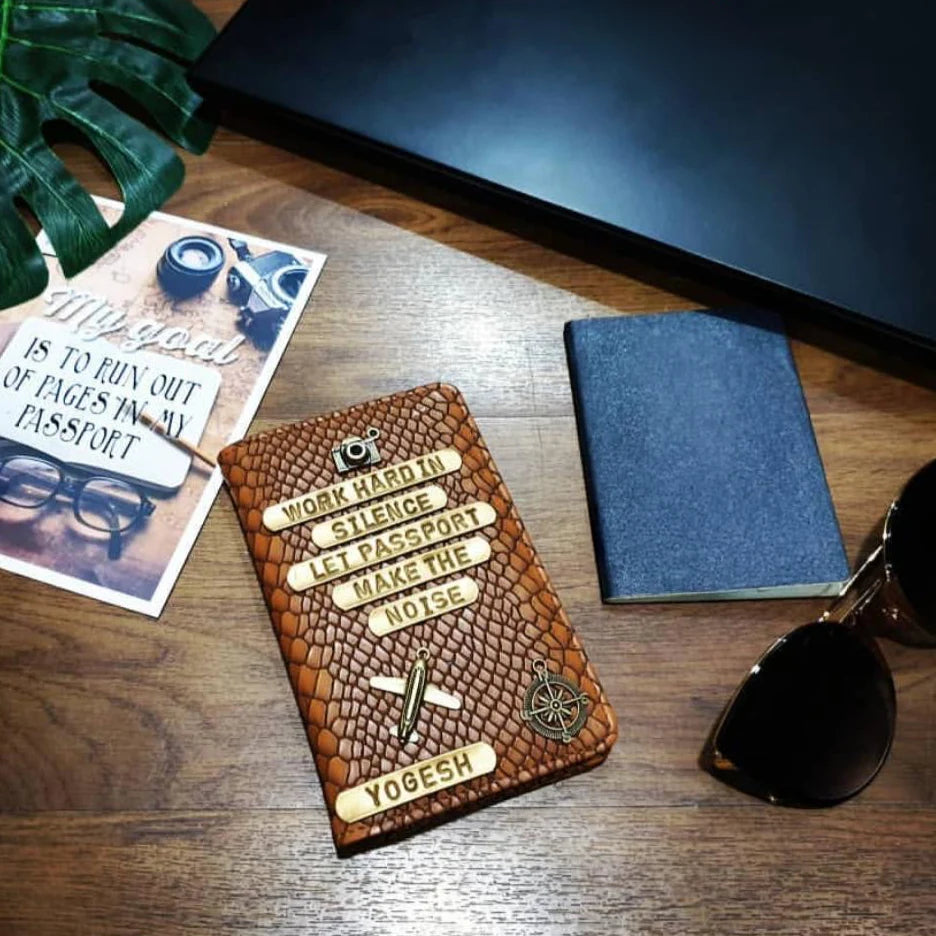 A textured brown leather passport cover with embossed text and travel-themed graphics, styled on a wooden tabletop with a blue notebook, sunglasses, a printed card, and a dark laptop in the background.