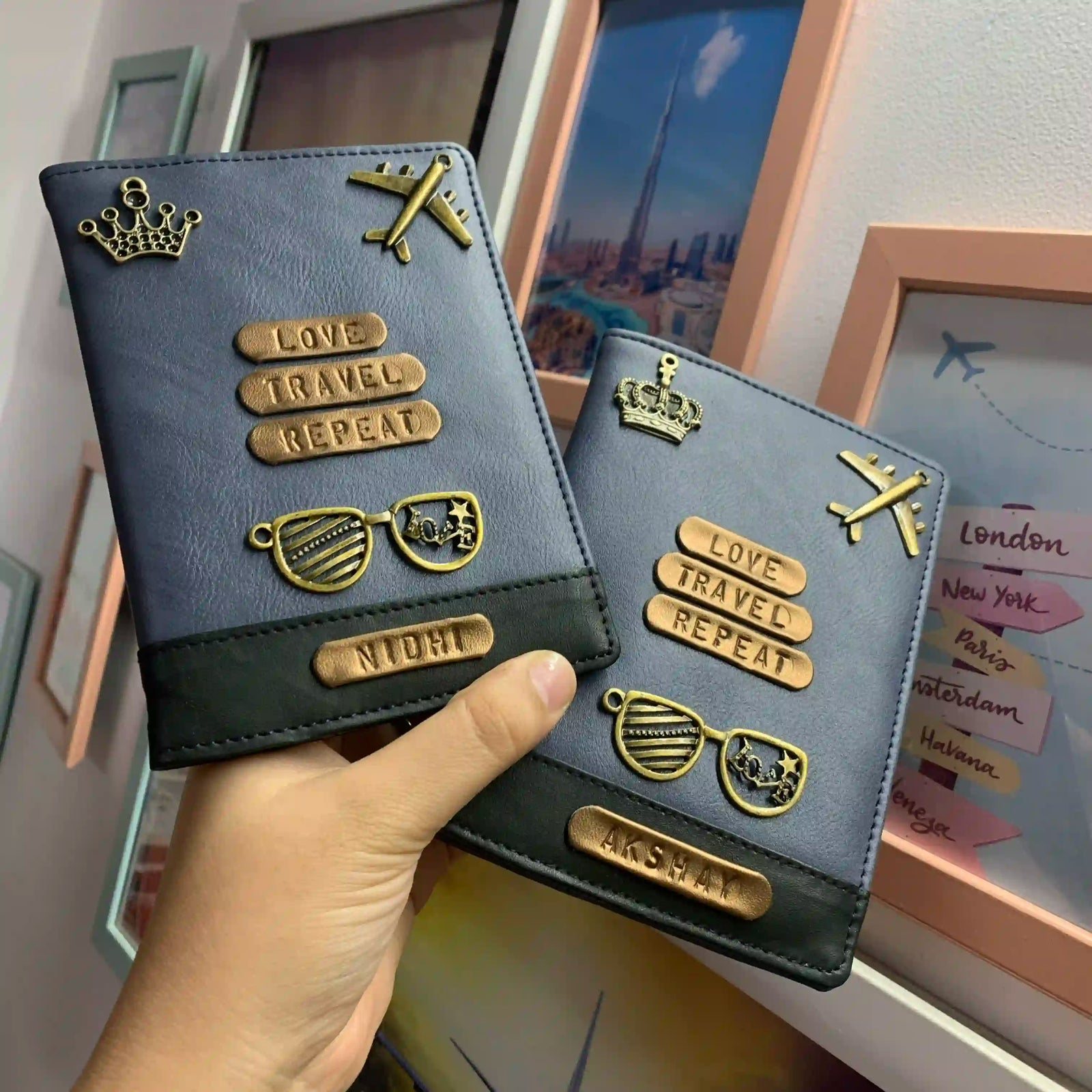 A hand holds two dark blue passport cover books decorated with gold-tone travel-themed charms, including a crown, airplane, sunglasses, and text plates reading 'LOVE TRAVEL REPEAT' and 'NIGHT'. The background shows framed travel posters and a wall- TPC Gifts