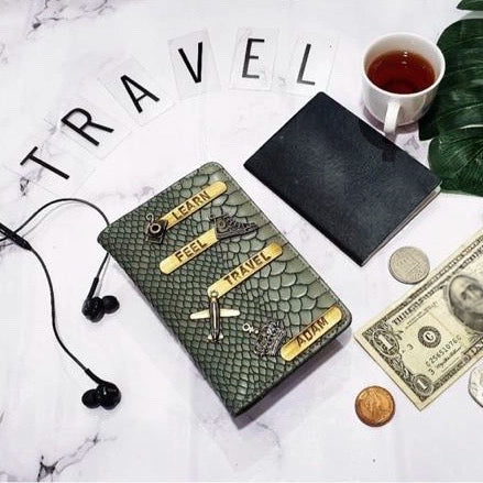 A textured grey  reptile-print travel passport cover with gold lettering and airplane embellishments, styled on a white marble surface with earbuds, a black notebook, a cup of tea, coins, a dollar bill, and the word 'TRAVEL' spelled out in letter tiles.
