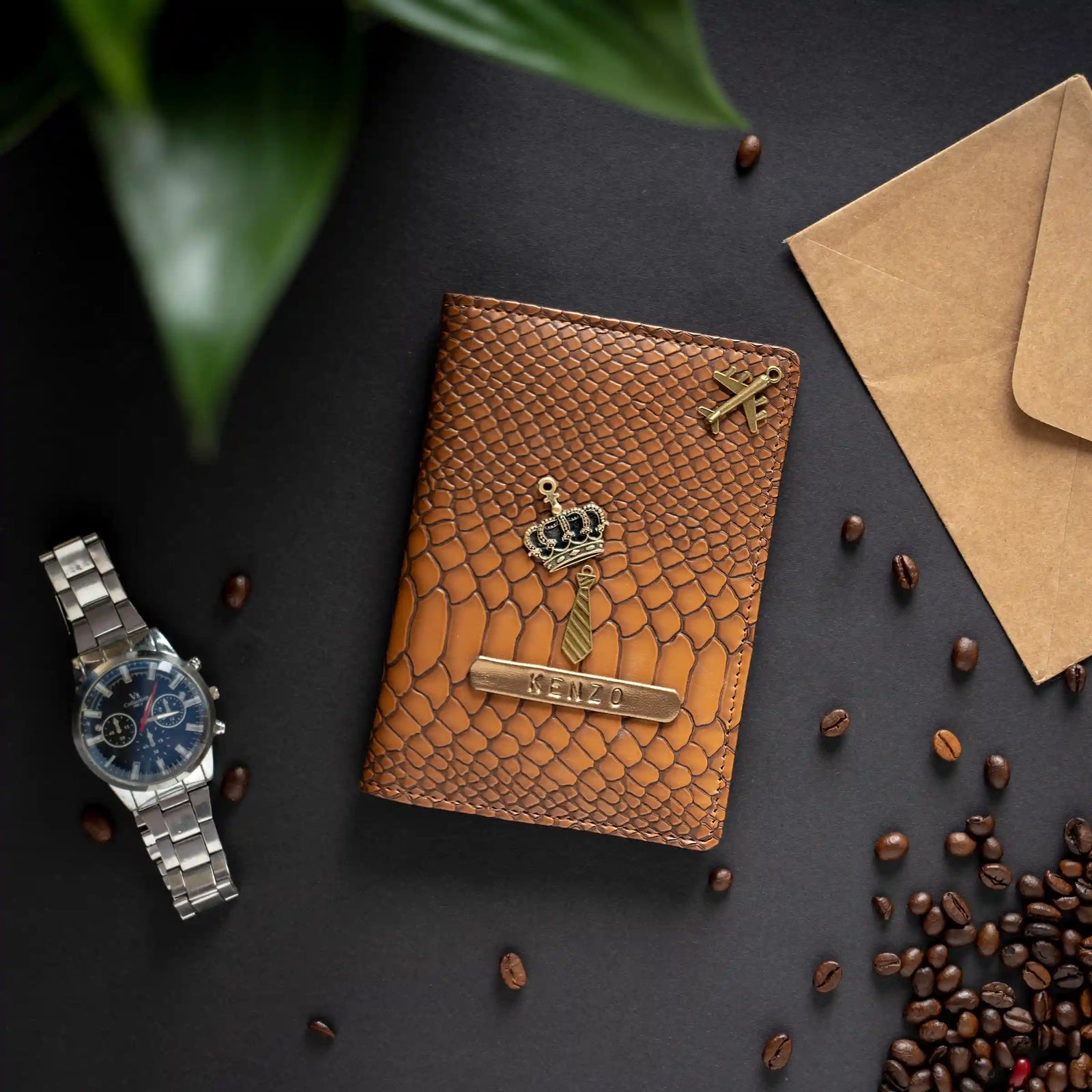 A brown snake embossed leather passport cover with a gold crown and nameplate, styled in a flat-lay with a silver watch, coffee beans, a kraft envelope, and green leaves on a dark background.