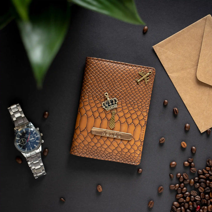 A brown snake embossed leather passport cover with a gold crown and nameplate, styled in a flat-lay with a silver watch, coffee beans, a kraft envelope, and green leaves on a dark background.