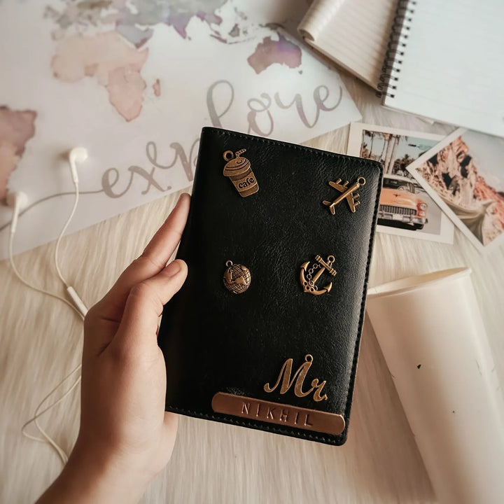 A hand holds a black leather passport cover decorated with gold-toned metal charms and a custom nameplate reading &