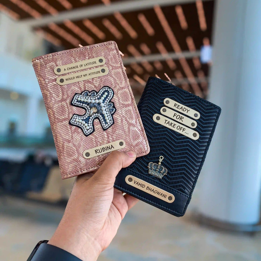 A hand holds two custom-designed passport covers in a blurred airport indoor setting. One passport cover is a textured pinkish-gold with a rhinestone plane and the other is black ziggy geuine leather passport cover with charm,quote and a name on it  - TPC Gifts 