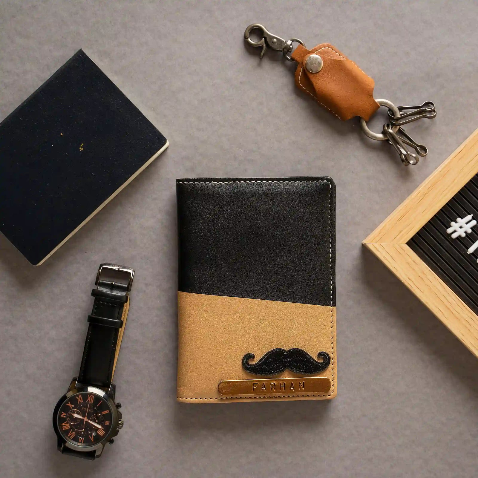 A slant  two-tone leather passport cover with a black top half and tan bottom half, featuring a black mustache graphic and a gold-toned nameplate reading 'RAFAEL'. The passport cover is styled in a flat-lay arrangement on a gray surface alongside a navy notebook, a wristwatch, a tan leather keychain, and a wooden letter board.
