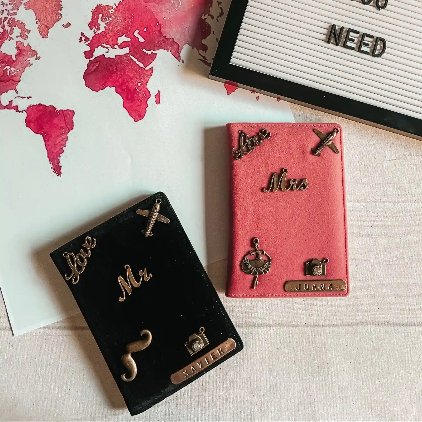 Two personalized passport covers, one black and one pink, decorated with metallic travel-themed charms and names, displayed on a light wood surface next to a world map print and a letter board- TPC Gifts