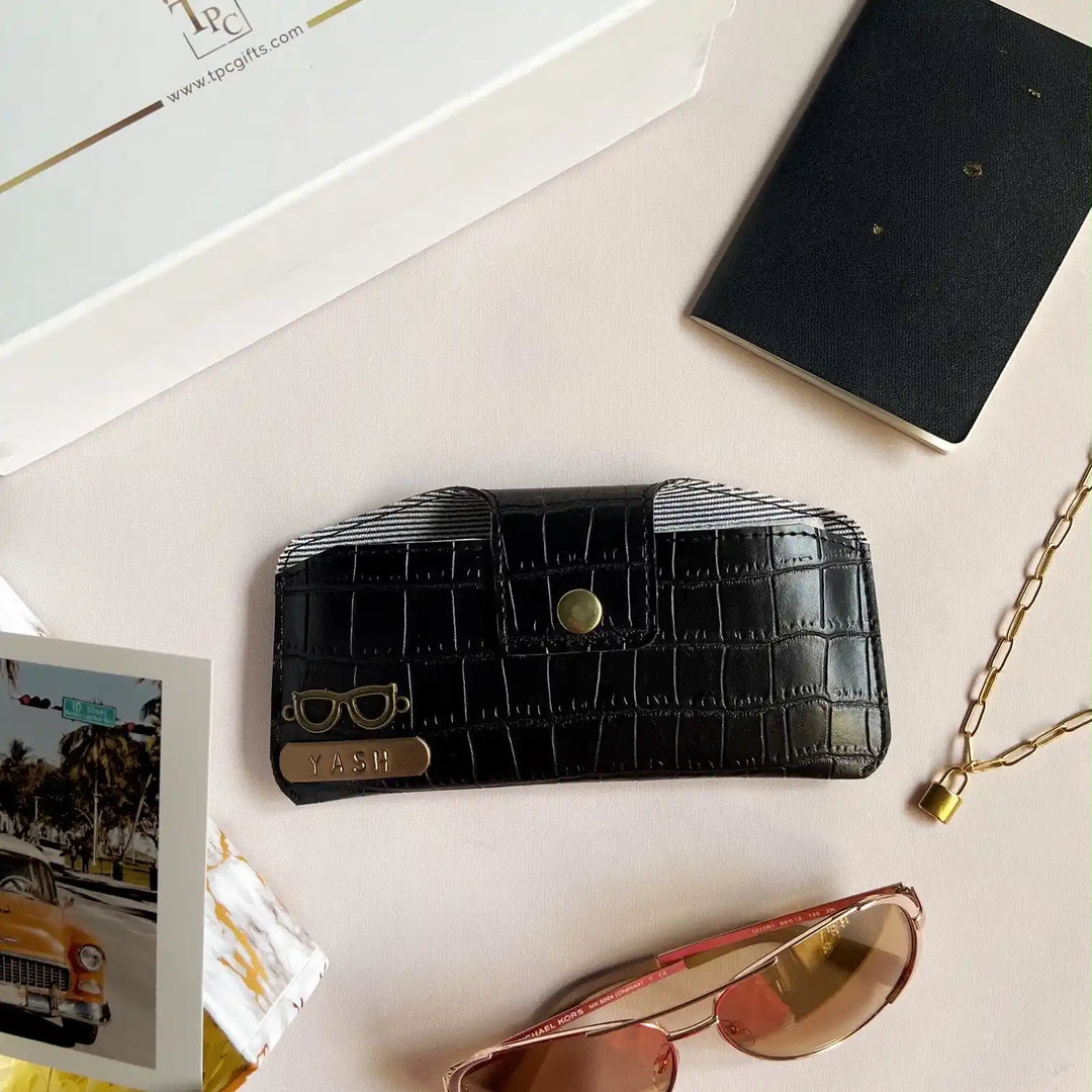 A black crocodile texture sunglass cover displayed alongside a passport, a book, and a pair of sunglasses on a light-colored surface.