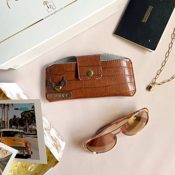 A brown crocodile texture sunglass cover displayed alongside a passport, a book, and a pair of sunglasses on a light-colored surface -TPC Gifts