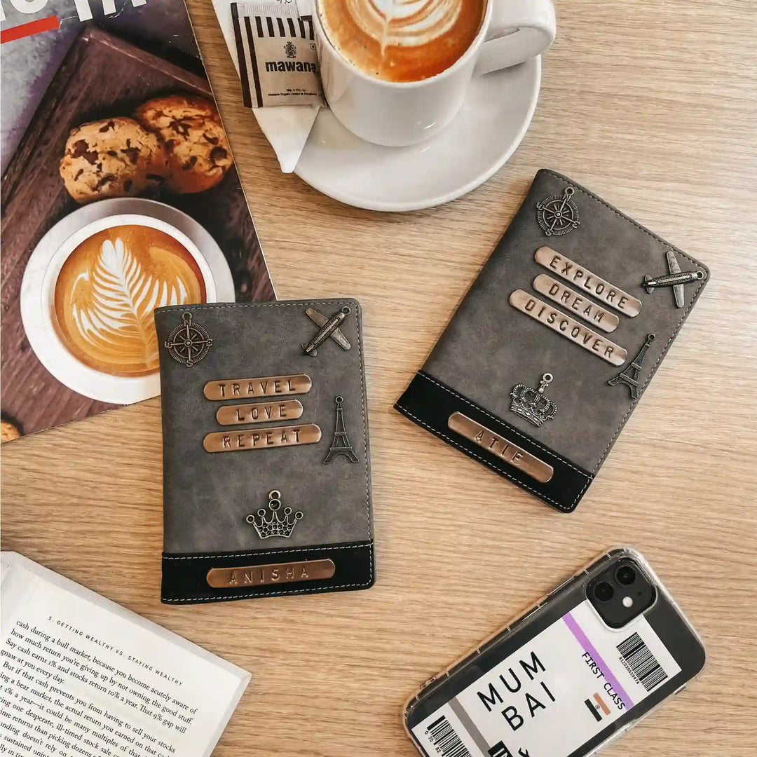 Two dark gray italian leather passport covers with travel-themed metal embellishments are displayed on a light wood tabletop alongside coffee cups, cookies, an open book, and a smartphone case.