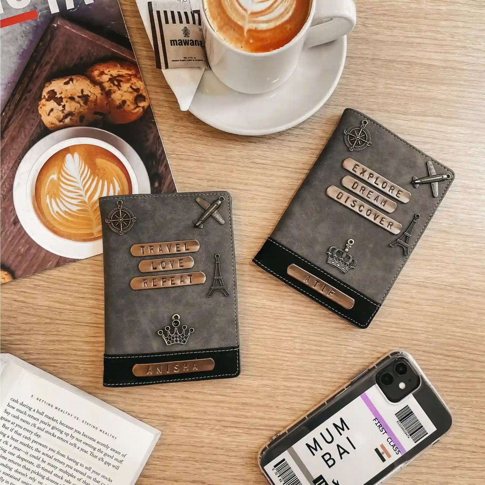Two dark gray italian leather passport covers with travel-themed metal embellishments are displayed on a light wood tabletop alongside coffee cups, cookies, an open book, and a smartphone case.