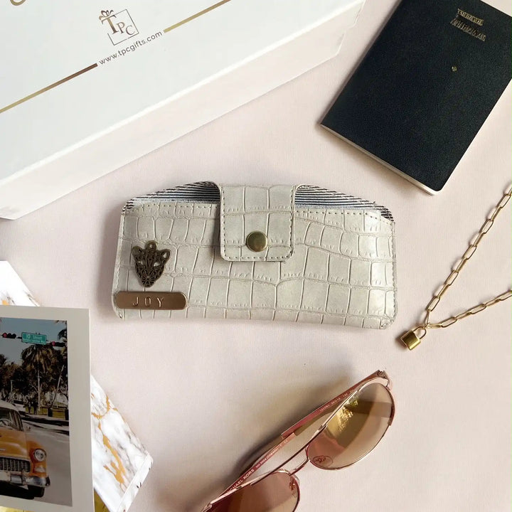 A grey crocodile texture sunglass cover displayed alongside a passport, a book, and a pair of sunglasses on a light-colored surface.