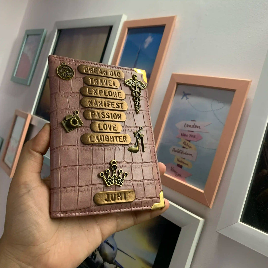 A hand holds a pink onion crocodile embossed wallet decorated with gold-toned metal charms and text plaques, set against a background of framed wall art.