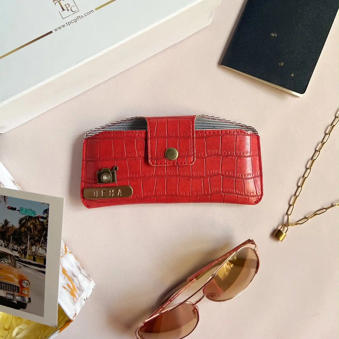 A red crocodile texture sunglass cover displayed alongside a passport, a book, and a pair of sunglasses on a light-colored surface -TPC Gifts