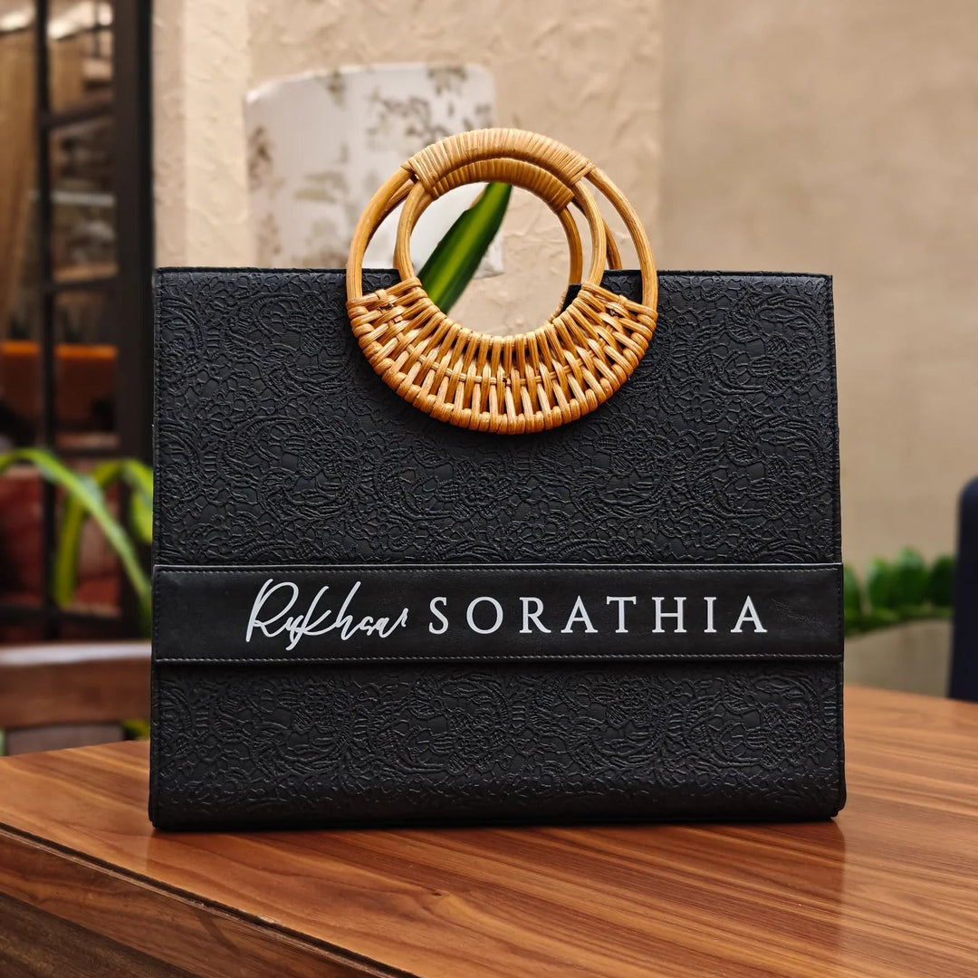 A black personalized tote bag with a cane handle, placed on a wooden table.