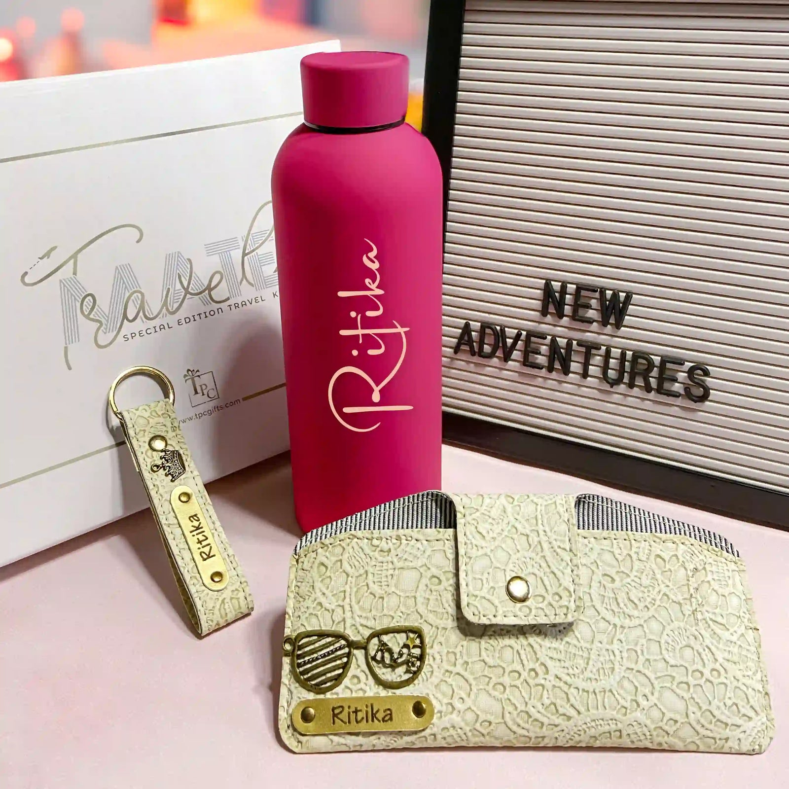 A luxury travel gift set including a pink custom water bottle, a beige sunglass cover, and a gold keychain with 'NEW ADVENTURES' sign displayed with branded packaging on a pink surface.