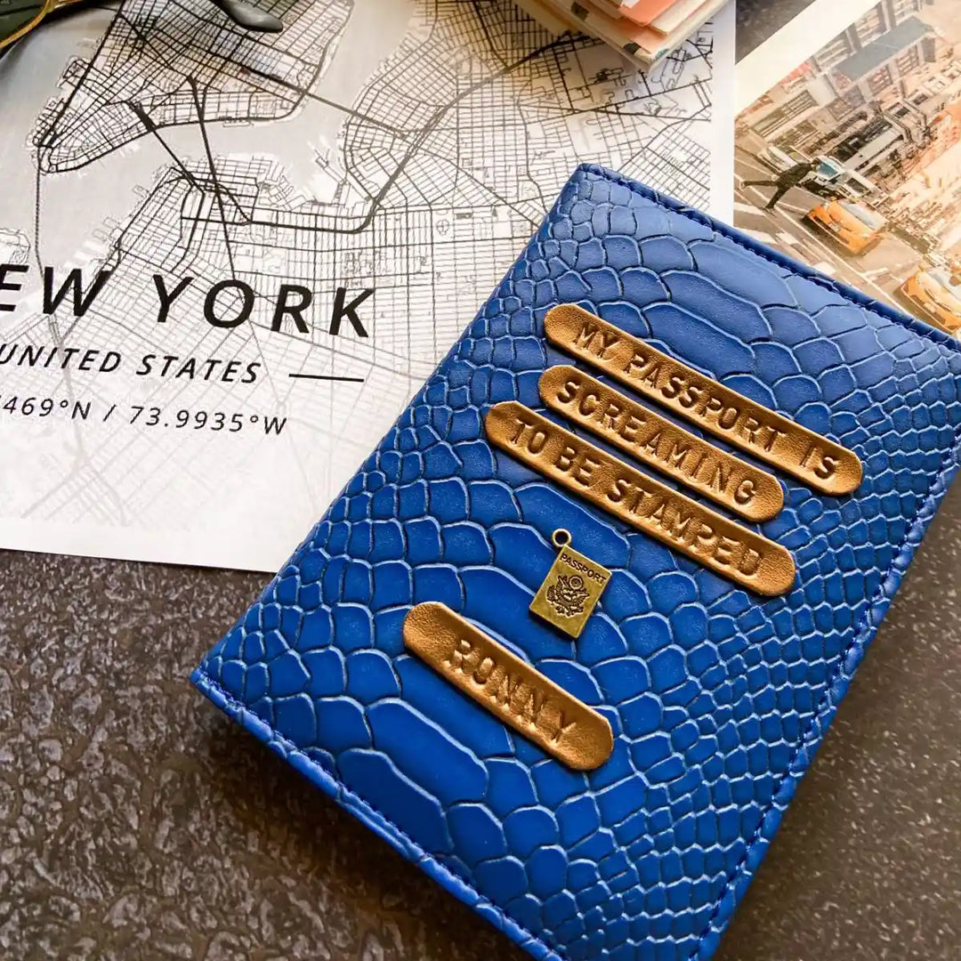 A blue snake textured passport cover featuring gold-toned nameplates and charms, styled on a dark tabletop next to a New York City map print.
