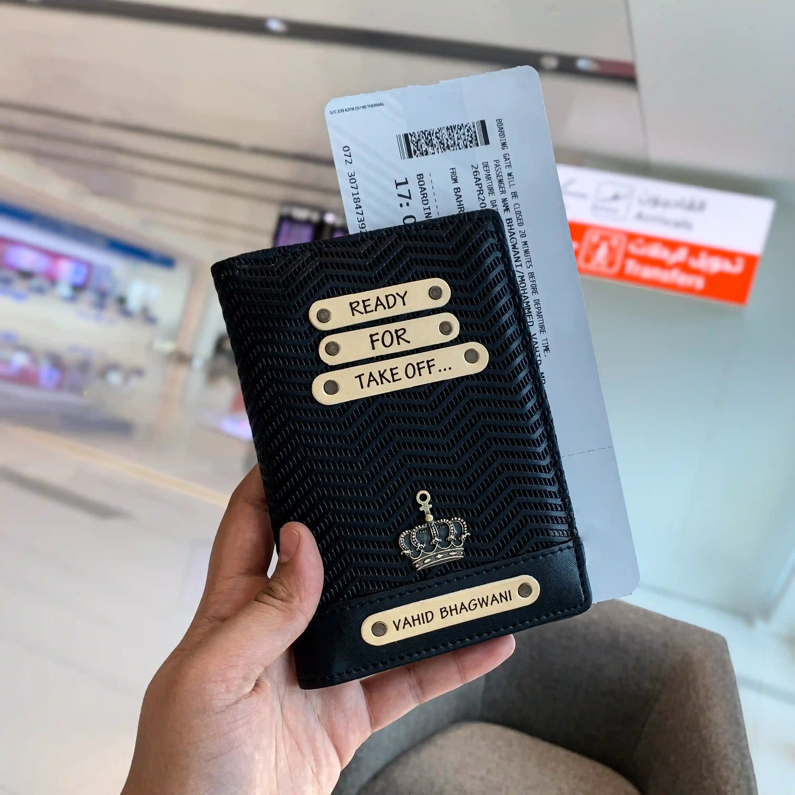A hand holding a black personalized passport cover with text on it, displayed in an airport setting from tpc gifts