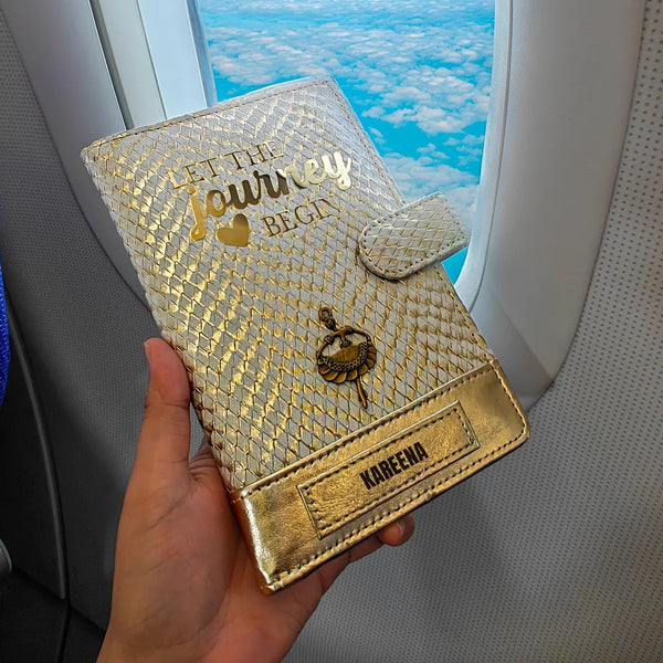 A personalized travel wallet by TPC Gifts with gold and ivory colors, featuring text and a crest design, held in a person's hand while on an airplane.