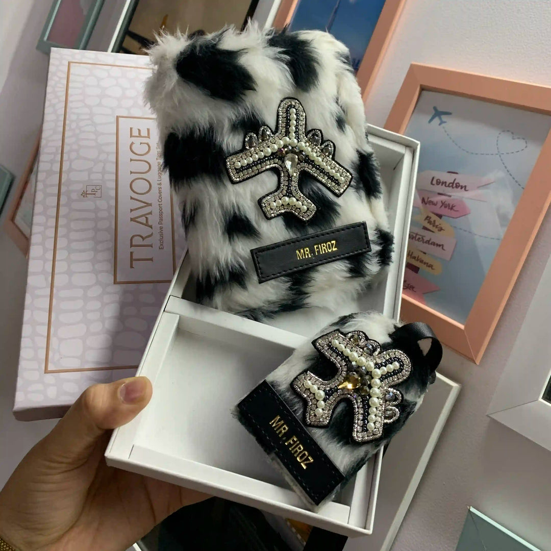 A hand holds an open white box displaying a black-and-white furry luggage tag and matching box lid. The tag has a rhinestone-embellished airplane emblem and a black label reading &