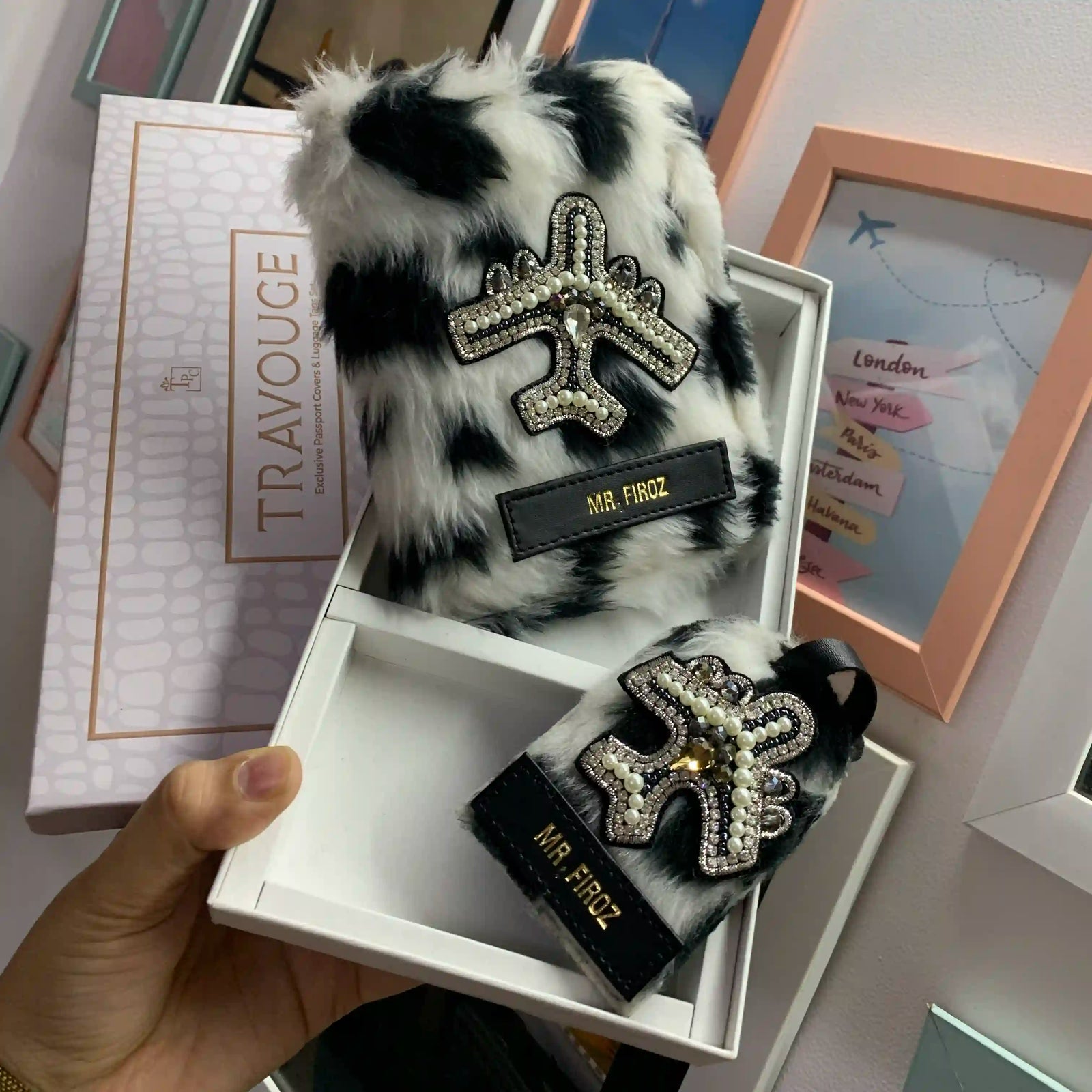 A hand holds an open white box displaying a black-and-white furry luggage tag and matching box lid. The tag has a rhinestone-embellished airplane emblem and a black label reading 'MR.