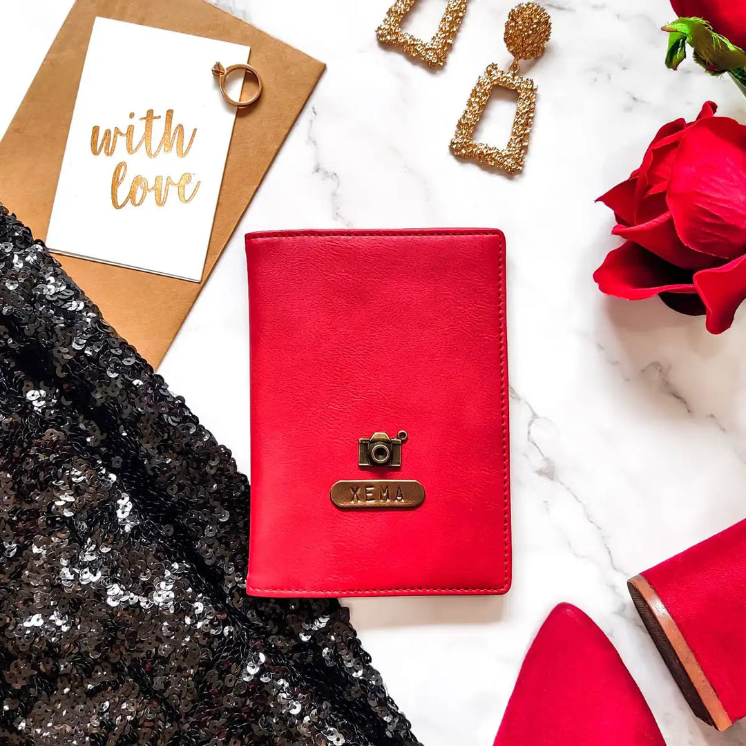 A red passport cover with a personalized name tag and charm, placed on a marble surface alongside a pair of earrings, a card, and a rose - TPC Gifts