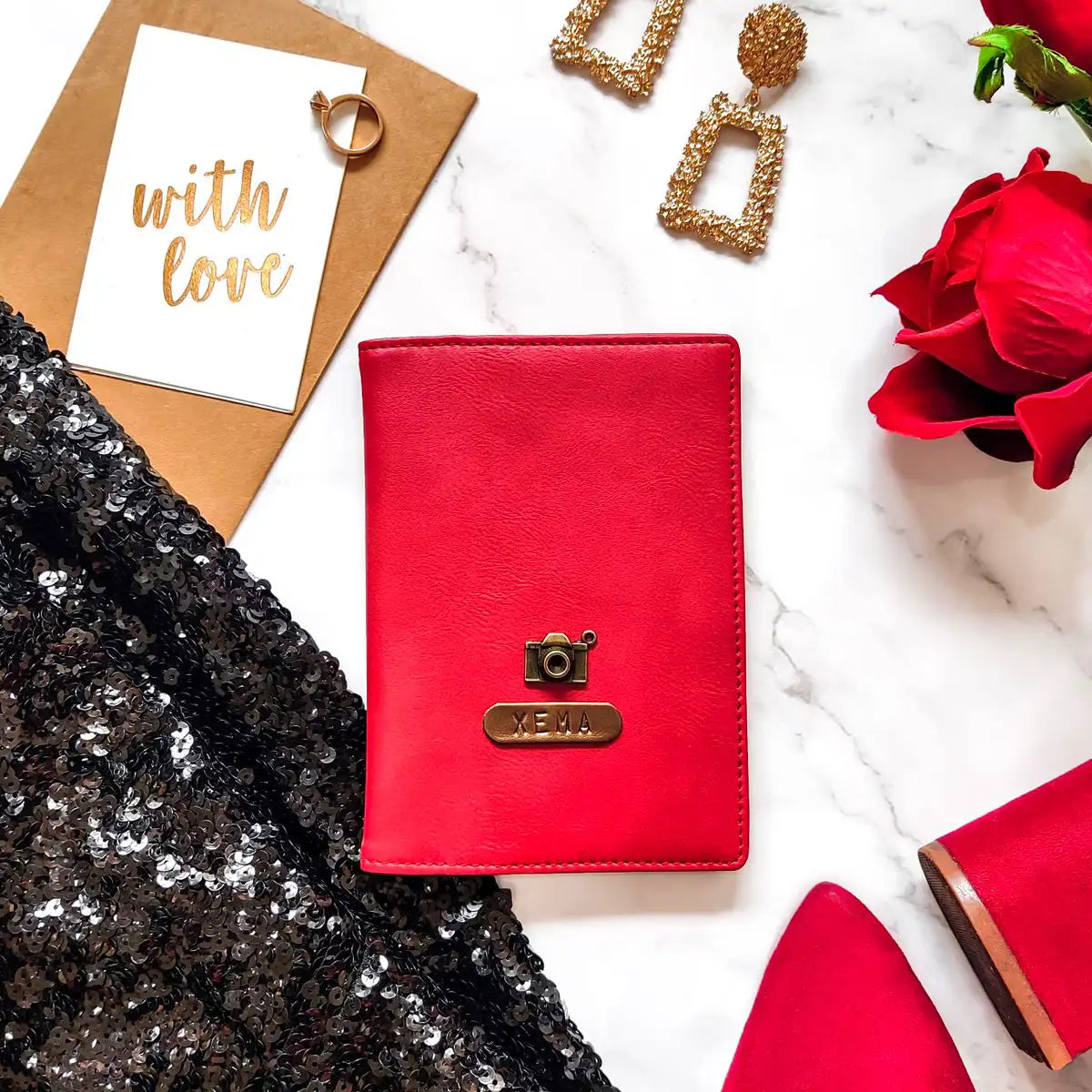 A red passport cover with a personalized name tag and charm, placed on a marble surface alongside a pair of earrings, a card, and a rose - TPC Gifts
