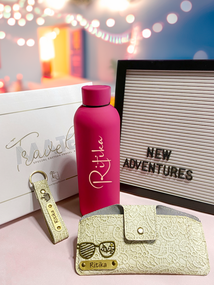 A luxury travel gift set including a pink custom water bottle, a beige sunglass cover, and a gold keychain, displayed against a backdrop with &