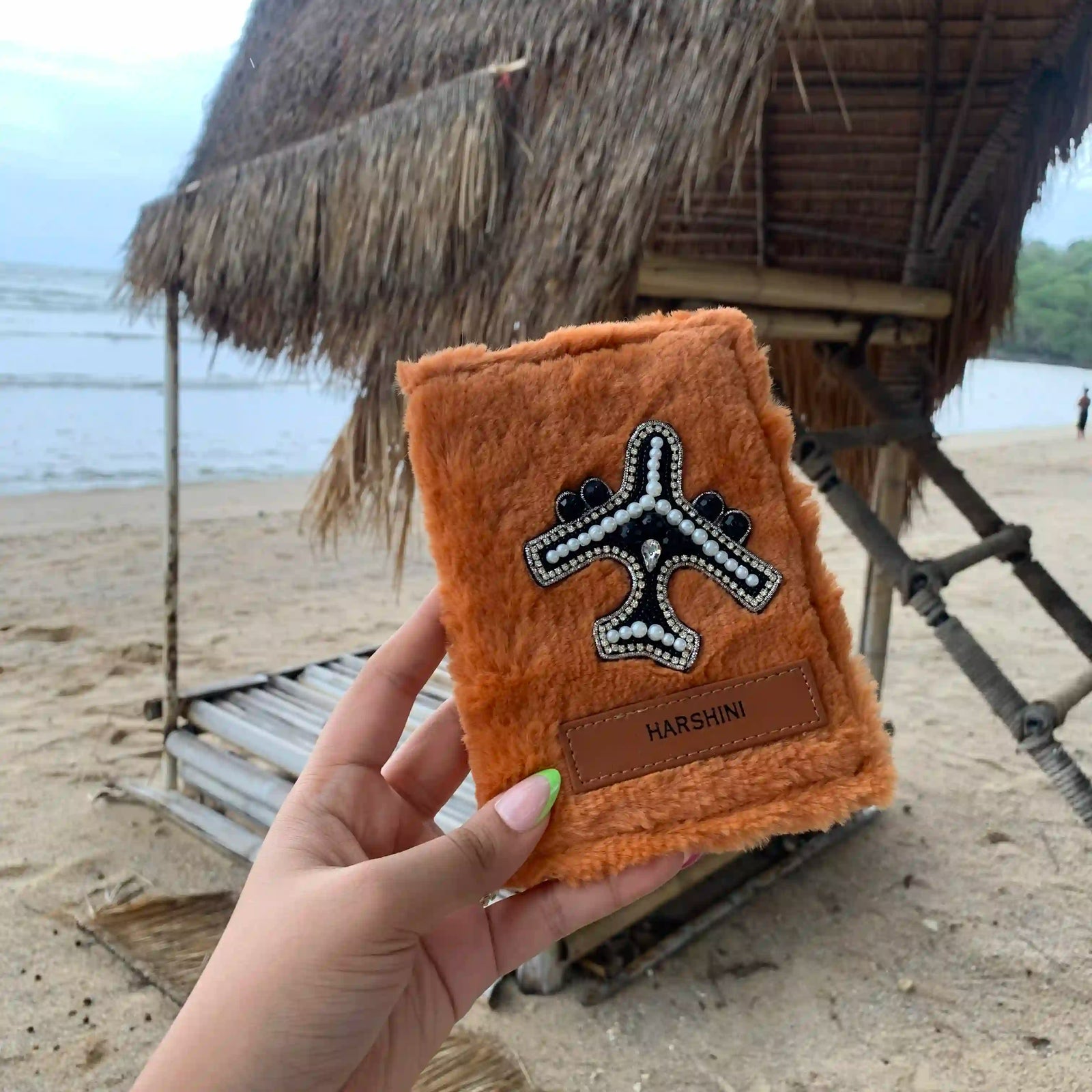 A hand holds a small orange-brown fuzzy pouch with a black-and-white airplane patch and a brown leather tab reading HARSHINI, set against a beach scene with a thatched structure in the background.
