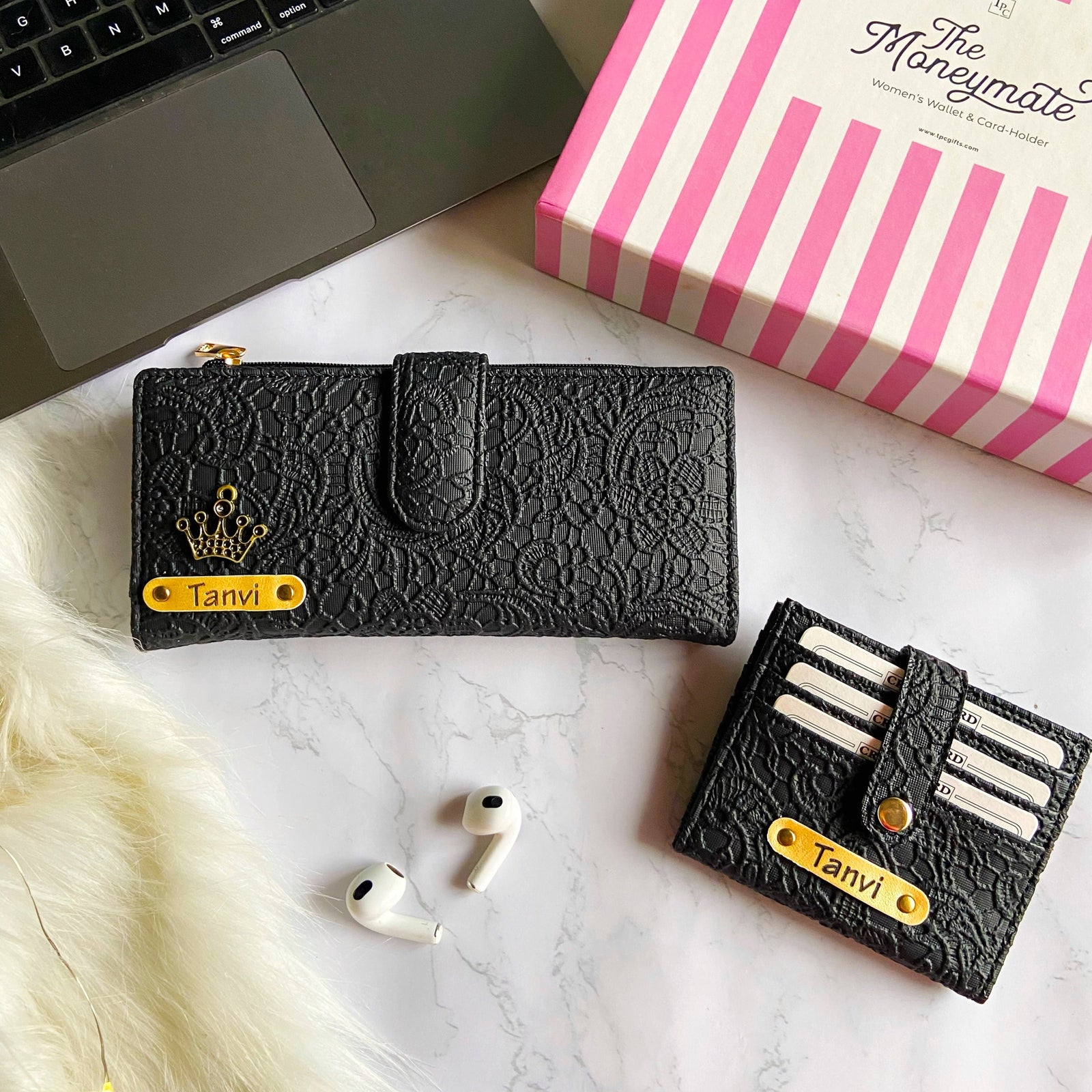 A black textured wallet and card holder with 'TANVI' printed on the front, alongside a pair of white earphones and a pink striped box in the background.