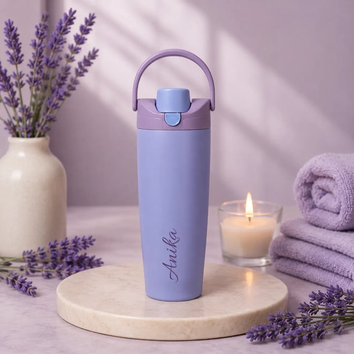 A lavender insulated water bottle with a matching carrying loop lid, standing on a round marble tray. The bottle has the name &