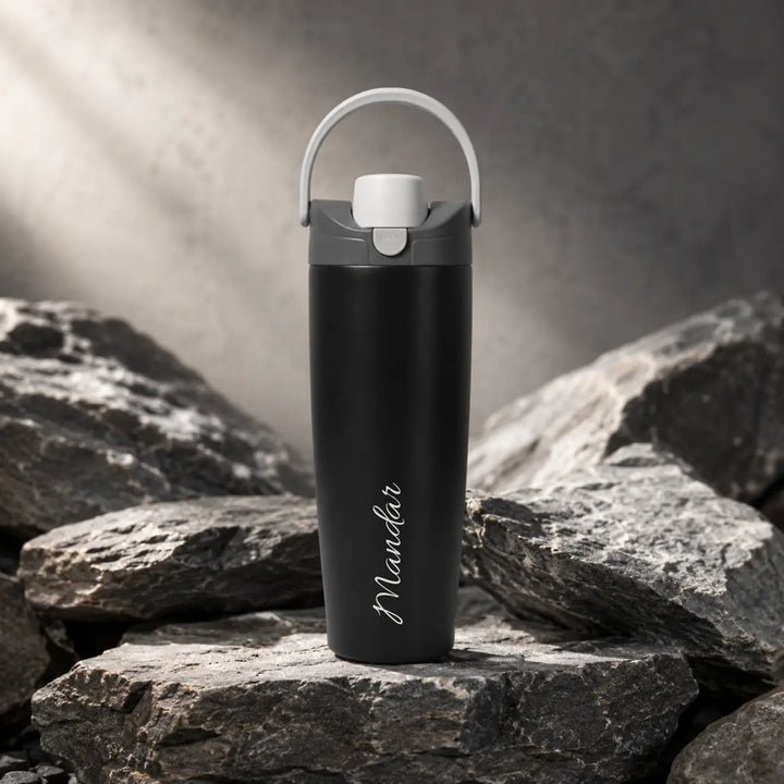 A matte black insulated water bottle with a white carrying loop and white lid, standing on dark rocks with a gray textured background- TPC Gifts