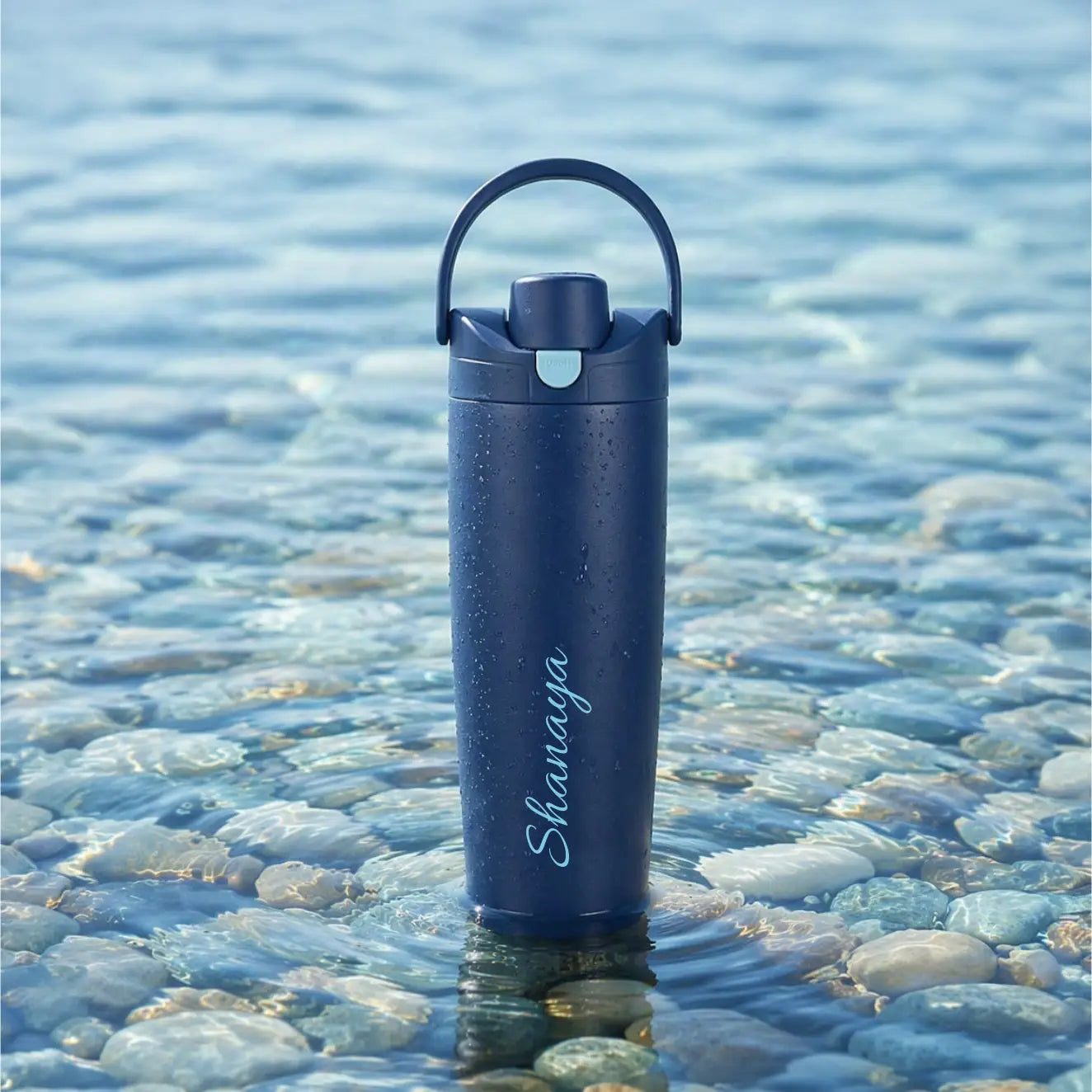 A dark navy blue insulated water bottle with a carrying loop and the brand name 'Shunaga' printed vertically, standing in shallow water over smooth blue-gray pebbles- TPC Gifts