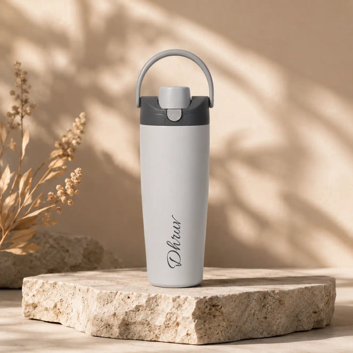 A light gray insulated water bottle with a dark gray lid and handle, standing on a textured stone slab against a warm beige background with soft leaf shadows. The bottle is personalized with the name &