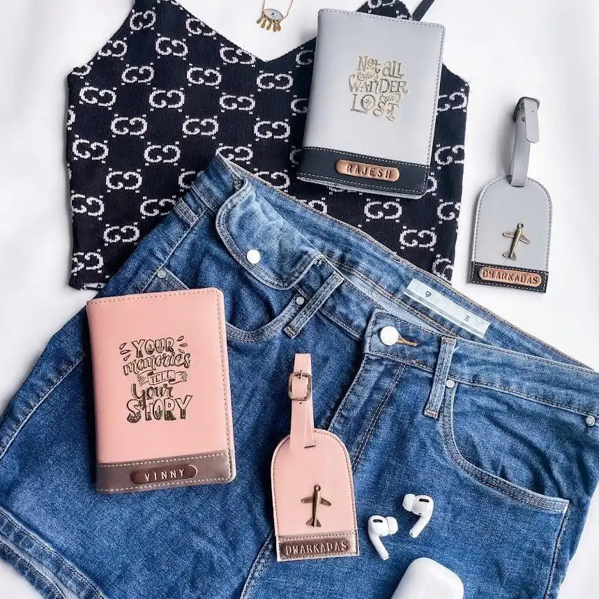A styled flat-lay of multiple faux leather travel accessories, including a black and white double-G monogram top, a gray passport holder with gold lettering, a pink passport holder, and matching luggage tags, arranged with blue denim shorts on a white background.