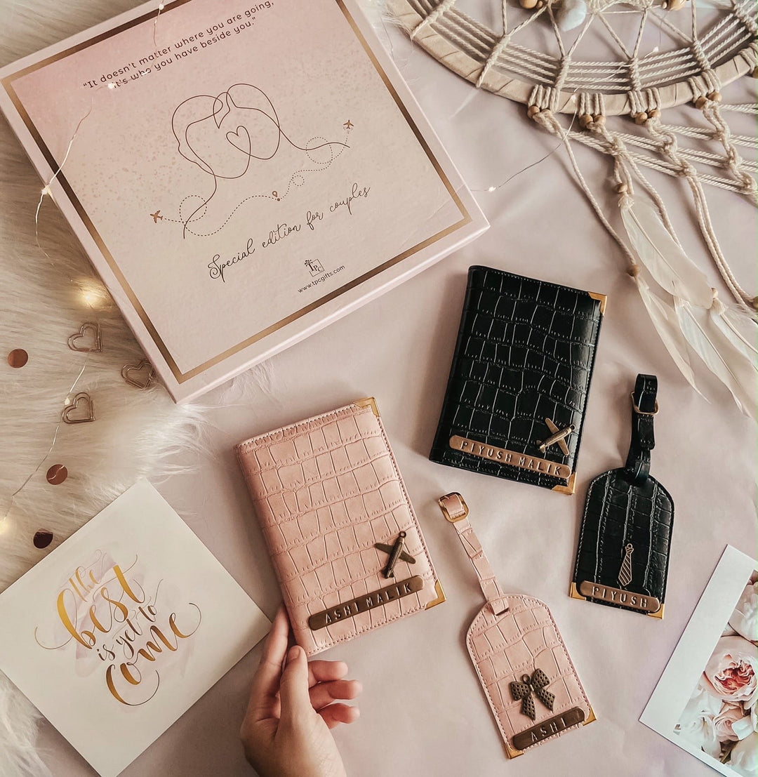 A set of couple passport covers and luggage tags in salmon pink and black croco texture displayed alongside a luxury gift box and personalized charms.