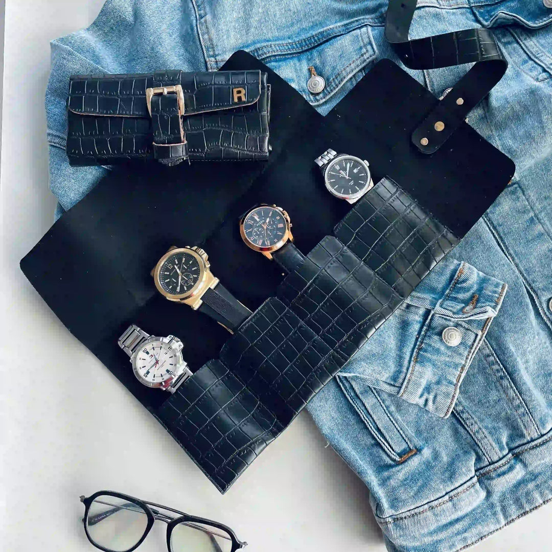 A black crocodile-embossed watch roll organizer laid flat on a light surface, holding several wristwatches, with a matching black watch pouch and a light blue denim jacket nearby. No person or real environment is present, so this is Product/Packaging rather than Lifestyle.