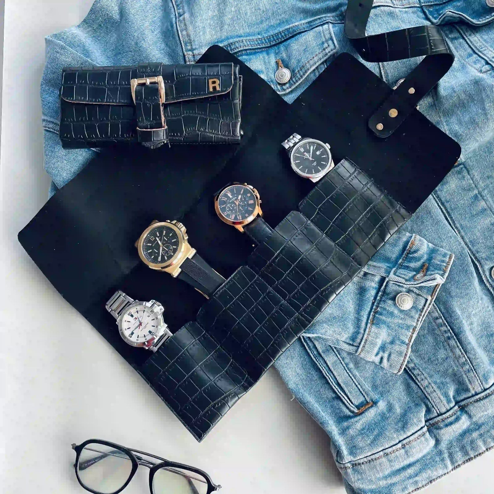 A black crocodile-embossed watch roll organizer laid flat on a light surface, holding several wristwatches, with a matching black watch pouch and a light blue denim jacket nearby. No person or real environment is present, so this is Product/Packaging rather than Lifestyle.